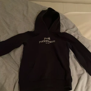 Svart hoodie från Peak Performance - Säljer en stilren svart hoodie från Peak Performance med vit logga på bröstet. Perfekt för kyliga dagar med sin mjuka och bekväma passform. Huvan ger extra värme och stil. Passar både till vardags och avslappnade tillfällen.
