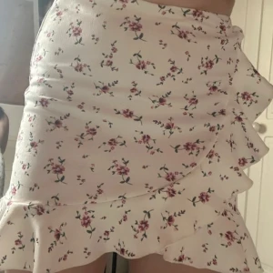 Blommig kjol från NLY One - Säljer en superfin blommig kjol från NLY One i storlek M. Den är vit med söta rosa blommor och har en volangdetalj längst ner. Perfekt för sommardagar eller en mysig vårutflykt. Kjolen är i bra skick och passar perfekt till en enkel topp eller blus. 🌸