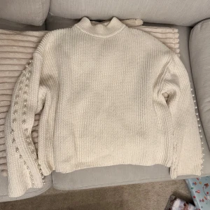 Vit stickad tröja med pärldetaljer - Säljer en stilren vit stickad tröja med pärldetaljer längs ärmarna. Tröjan har en hög krage och långa ärmar, perfekt för kyligare dagar. Den är både mysig och elegant, vilket gör den till ett mångsidigt tillskott i garderoben.