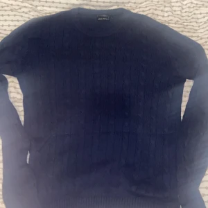 Mörkblå stickad tröja från East West - Säljer en snygg mörkblå stickad tröja från East West. Perfekt för kyliga dagar med sitt klassiska kabelstickade mönster. Tröjan har långa ärmar och en rund halsringning. Passar både till vardags och lite finare tillfällen. 🧶💙