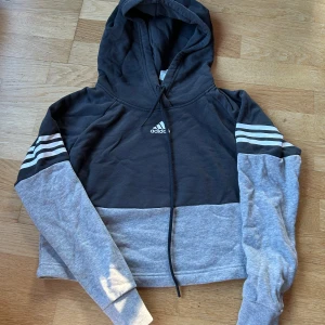 Grå och svart hoodie från Adidas - Säljer en snygg och bekväm kort hoodie från Adidas i grått och svart. Den har de klassiska tre ränderna på ärmarna och en huva. Perfekt för en avslappnad stil eller träning. Den är i mycket bra skick och använd ett fåtal gånger! 🖤