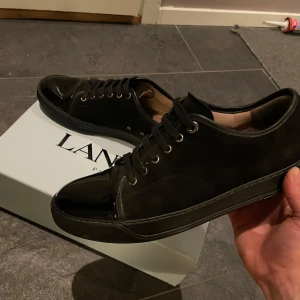 Svarta sneakers från Lanvin 9/10 skick - Säljer ett par stilrena svarta sneakers från Lanvin i mycket bra skick och har en snygg glansig finish. Nypris 5000 kr. De har klassisk snörning och kommer med originalförpackning, dushbags, extra snören och skovård från Jason Markk. Perfekta för både vardag och fest! 
