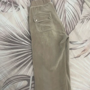 Beige mjukisbyxor från Juicy Couture - Säljer ett par superbekväma beige mjukisbyxor från Juicy Couture. De har en hög midja och är gjorda i mjuk velour. Byxorna har praktiska fickor med knappar och en avslappnad passform. Perfekta för mysiga dagar hemma eller en avslappnad dag ute.