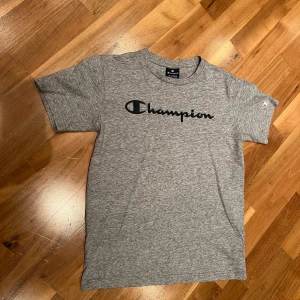 Grå t-shirt från Champion - Säljer en snygg grå t-shirt från Champion i storlek 162/167 (barnL). Den är i mycket bra skick och har en klassisk Champion-logga tryckt i svart på bröstet. Perfekt för vardagsbruk eller träning. Materialet känns mjukt och bekvämt. Passa på att köpa denna stilrena t-shirt!