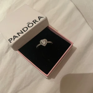 Silverring med hjärtformad sten från Pandora - Säljer en elegant silverring från Pandora med en glittrig hjärtformad sten i mitten. Ringen har små stenar längs bandet som ger extra bling. Perfekt för speciella tillfällen eller som en fin present. Kommer i originalförpackning.