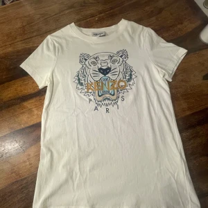 Vit t-shirt med tigertryck från Kenzo - Säljer en snygg vit t-shirt från Kenzo i storlek Xs som är väldigt liten i storleken. Den har ett coolt tigertryck i mitten med texten 'Kenzo Paris' i guld och mörkblått. Perfekt för en casual look eller att styla upp med en kavaj. T-shirten är i bomull och har korta ärmar. Passar perfekt till både jeans och kjol!