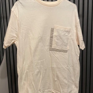 Beige t-shirt med fickdetalj från Another Influence - Säljer en stilren beige t-shirt från Another Influence i storlek XS. Den är gjord av mjuk bomull och har en unik fickdetalj på bröstet med en extra kant i en ljusare nyans. Perfekt för en avslappnad vardagslook! (Free size)