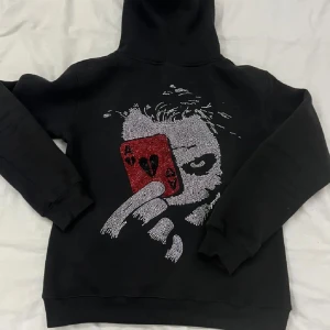 RHINESTONE zip hoodie - Cool svart hoodie med ett glittrigt motiv av en person som håller ett rött kort med hjärter. Framsidan har texten 'Masterme' i gotisk stil. Perfekt för höst och vinter, med dragkedja och fickor framtill.