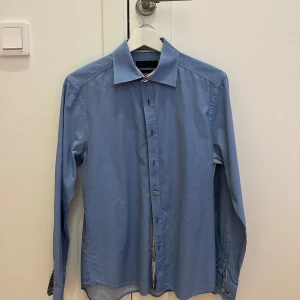 Blå skjorta från Riley - Säljer en snygg blå skjorta från Riley i regular fit. Skjortan har långa ärmar och knappar framtill. Perfekt för både vardag och festliga tillfällen. Den är i bra skick och gjord av ett bekvämt material som känns skönt mot huden.