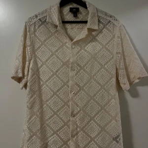 Beige spetsmönstrad skjorta från H&M - Säljer en snygg beige skjorta från H&M i regular fit. Skjortan har ett unikt spetsmönster och korta ärmar, perfekt för sommardagar. Den har knappar framtill och en klassisk krage. Passar både till vardags och festliga tillfällen. 🕶️