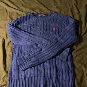 Blå stickad tröja från Polo Ralph Lauren - Säljer en snygg blå stickad tröja från Polo Ralph Lauren med klassiskt flätmönster. Den har en liten röd logga på bröstet och ribbade muddar vid ärmslut och nederkant. Perfekt för kyliga dagar! 