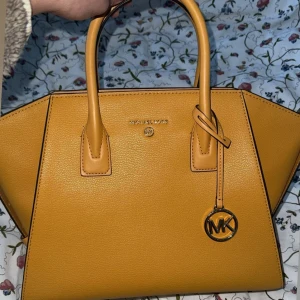 Gul handväska från Michael Kors - Säljer en snygg gul handväska från Michael Kors i skinn. Väskan har en stilren design med två handtag och en dekorativ MK-berlock. Perfekt för att liva upp vilken outfit som helst och perfekt för sommaren och hösten! Den är aldrig använd och kommer med dustbag och äktahetskort i väskan. Priset kan diskuteras vid snabb affär