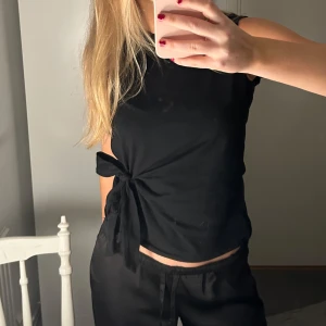 Svart ärmlös topp med knyt - Snygg svart ärmlös topp i T-shirt material med knytning i sidan. Till vardag och fest! Köpt från Sellpy okänt märke men kan kolla upp. Toppen är lite nopprig på insidan💗