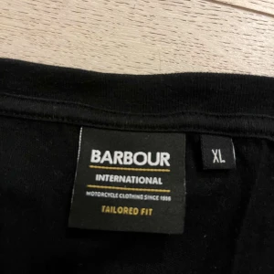 Svart t-shirt från Barbour International - Säljer en svart t-shirt från Barbour International i storlek XL. Den har en klassisk design med korta ärmar och en gul logga på ärmen. Perfekt för en avslappnad stil eller som basplagg i garderoben.