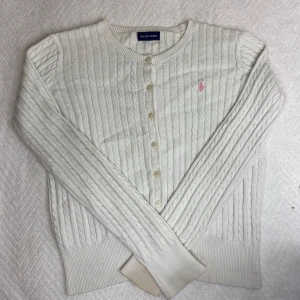 Vit kabelstickad kofta från Ralph Lauren - Säljer en superfin vit kabelstickad kofta från Ralph Lauren. Den har långa ärmar och knappar framtill. Detaljerad med en rosa broderad logga på bröstet. Köpte för 2295 kr. Passar S!💕