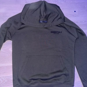 Olivgrön hoodie från Essentials - Säljer en olivgrön hoodie från Essentials Fear of God. Den har en klassisk design med känguruficka och tryck både fram och bak. Perfekt för en avslappnad stil och passar bra till höst och vår. Skön och stilren, ett måste i garderoben!