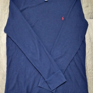 Ralph Lauren Tröja  - Säljer en snygg blå tröja från Polo Ralph Lauren. Den har en diskret ribbad struktur och det klassiska röda broderade logotypen på bröstet. Perfekt för både vardag och lite finare tillfällen. Passar bra till jeans eller chinos!