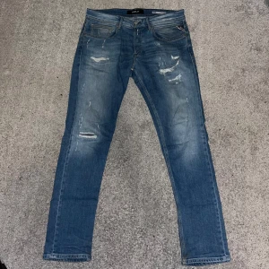 Blå jeans från Replay - Säljer ett par snygga blå jeans från Replay i storlek 31. De har en cool slitningseffekt och en klassisk femficksdesign. Perfekta för en avslappnad stil och passar till både vardag och fest. Dragkedja och knapp framtill för enkel stängning.