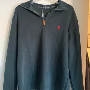Mörkgrön tröja från Ralph Lauren - Säljer en snygg mörkgrön tröja från Ralph Lauren i mycket bra skick. Den har en dragkedja vid halsen och det klassiska röda logotypbroderiet på bröstet. Perfekt för både höst och vår. Passar bra till både vardag och lite finare tillfällen.