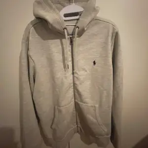 Säljer en stilren grå hoodie från Ralph Lauren med dragkedja och klassisk logga på bröstet. Perfekt för både höst och vår. Den har en bekväm passform och är tillverkad i mjukt material. Huvan har justerbara snören och det finns praktiska fickor framtill. Perfekt för en avslappnad look!