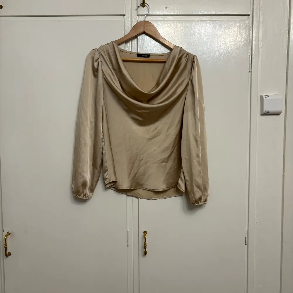 Snygg beige blus från Boohoo i ett glansigt silke material. Den har långa ärmar och en draperad halsringning som ger en elegant touch. Perfekt för både vardag och festliga tillfällen. Blusen är i jätte bra skick och redo för en ny ägare! Blusen har använts 4 gånger. . Toppar.