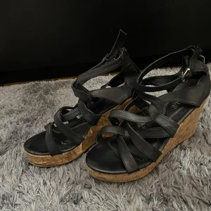 Svarta kilklackssandaler från Avenue - Snygga svarta sandaler med kilklack från Avenue. De har en korkliknande sula och flera remmar som ger en cool look. Perfekta för sommaren och festliga tillfällen. Dragkedja baktill för enkel på- och avtagning.
