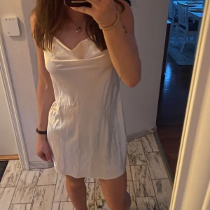 Vit sidenklänning - Säljer en elegant vit sidenklänning i kort modell. Klänningen är öppen i ryggen och har en vacker glansig yta som passar perfekt för sommarens fester eller skolavslutning. Den är i nyskick och ger en lyxig känsla. Perfekt för en somrig look!