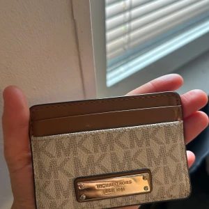 Kortfodral från Michael Kors - Säljer ett stilrent kortfodral från Michael Kors i beige och brunt. Det har ett snyggt mönster med märkets logga och en metallplatta framtill. Perfekt för att hålla dina kort organiserade och lättillgängliga. Passar både till vardags och fest!