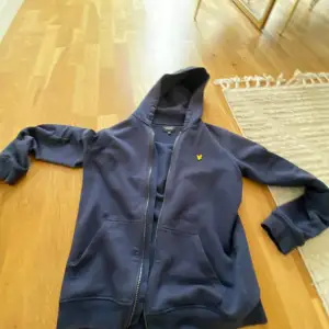 CHECKA MITT KONTO FÖR ATT HITTA SNYGGA KLOCKOR GANT RALPH LOUREN OCH MER .En klassisk mörkblå Lyle & Scott-hoodie med dragkedja. Den har den typiska broderade loggan i gult på bröstet och en enkel, tidlös design som passar bra till både avslappnade och mer uppklädda stilar. Materialet verkar vara mjukt och bra skick 