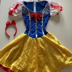 Snövit-inspirerad klänning med diadem - Säljer en superfin Snövit-inspirerad klänning i blått, gult och rött. Klänningen har en kort kjol och ärmlös design med röda banddetaljer. Perfekt för halloween, maskerad eller temafest! Inkluderar ett matchande rött diadem med rosett. Dragkedja bak för enkel påklädning. Ett av de röda banden på ena ärmen är trasigt.
