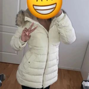 Vit pufferjacka med pälsdetalj - Säljer en supermysig vit pufferjacka med pälsdetalj på huvan. Perfekt för kalla vinterdagar! Jackan har en dragkedja framtill och är långärmad. Den är i mycket bra skick och håller dig varm och stilren hela säsongen.