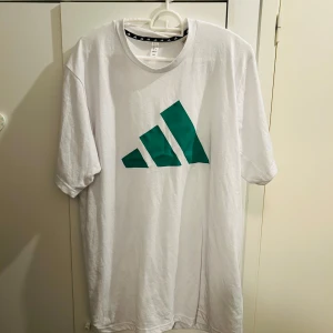 Vit t-shirt från Adidas - Säljer en vit t-shirt från Adidas i storlek L. Den har en stor grön logga på framsidan och är gjord i mjuk bomull. Perfekt för en avslappnad stil eller träning. T-shirten är kortärmad och har en normal passform. Skicka ett meddelande vid intresse!