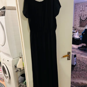 Svart långklänning - Elegant svart långklänning i ett klassiskt snitt. Perfekt för festliga tillfällen eller en stilren vardagslook. Klänningen har en rund hals och korta ärmar, vilket ger en tidlös och sofistikerad stil. Materialet ser ut att vara mjukt och bekvämt, vilket gör den lätt att bära hela dagen.