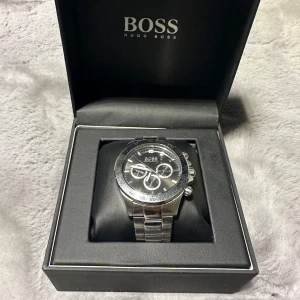 Hugo Boss klocka  - Med en boett och armband i rostfritt stål ger den en modern, elegant look. Urtavlan har tydliga index och en minimalistisk design som gör den lätt att läsa. Vattentålig och ergonomisk. Tillbehör, papper finns med och lådan  