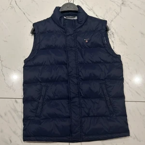 Mörkblå dunväst från GANT - Säljer en snygg mörkblå dunväst från GANT. Perfekt för höst och vår med sitt quiltade mönster och dragkedja framtill. Västen har en stilren design med GANT-logga på bröstet och randigt foder inuti. Passar både till vardags och lite finare tillfällen. Storlek för ungdomar 13-14 år.