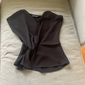 Svart omlottopp - Säljer en stilren svart omlottopp med korta ärmar från zara Perfekt för både vardag och fest! Den har en knytning i midjan som ger en smickrande passform. Toppen är i ett mjukt och bekvämt material.