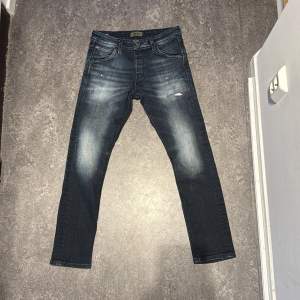 Säljer ett par snygga mörkblå jeans från Jack & Jones. De har en slim fit och är i mycket bra skick. Jeansen har en klassisk femficksdesign och en lätt slitning på framsidan för en cool look. Minimal fläck på båda knän. Annars fantastiskt skick!