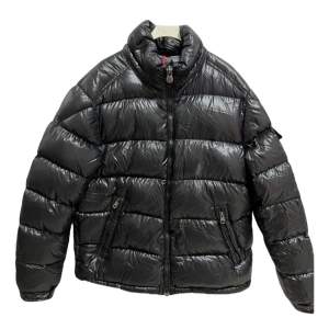 Säljer en snygg svart pufferjacka från Moncler i nyskick. Jackan är perfekt för vintern med sitt glansiga polyester-material och varma dunfyllning. Den har en dragkedja framtill och två praktiska fickor med dragkedjor. Moncler-loggan sitter på ärmen. Perfekt för kalla dagar! ❄️ Jackan är i storlek M, hör gärna av dig vid fler frågor!