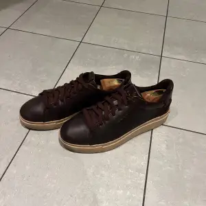 Säljer ett par snygga bruna sneakers från Gant i skinn. De har en stilren design med snörning och en beige sula med röd undersida. Perfekta för både vardag och festliga tillfällen. Skorna är i mycket bra skick och redo för en ny ägare!