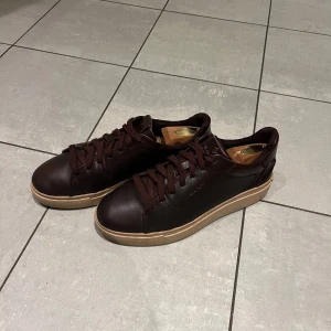 Bruna sneakers från Gant - Säljer ett par snygga bruna sneakers från Gant i skinn. De har en stilren design med snörning och en beige sula med röd undersida. Perfekta för både vardag och festliga tillfällen. Skorna är i mycket bra skick och redo för en ny ägare!