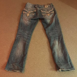 Rock Revival jeans med slitningar - Säljer ett par coola Rock Revival jeans i stl 31, de passar stl s-m. De är blå med snygga slitningar och har unika detaljer på bakfickorna.