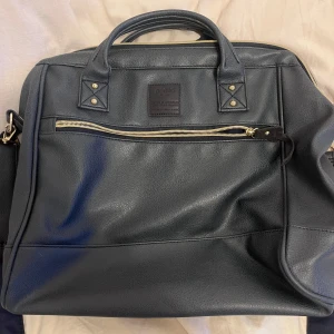 Svart datorväska med dragkedja - Snygg och praktisk svart datorväska i skinn med dragkedja framtill. Perfekt för att bära med sig datorn till jobbet eller skolan. Väskan har både handtag och en justerbar axelrem för bekväm användning. Den är i mycket bra skick och redo för nya äventyr!