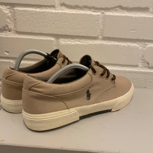 Beige sneakers från Polo Ralph Lauren - Snygga beige sneakers från Polo Ralph Lauren i mycket bra skick. De har en klassisk design med snörning och en vit sula. Perfekta för vardagsbruk och ger en stilren look. Märkets ikoniska logga syns på sidan av skorna.
