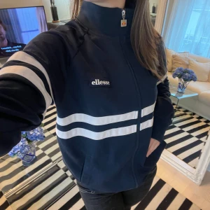 Ellesse zipper  - Säljer en snygg mörkblå jacka från Ellesse med vita ränder över bröstet och ärmarna. Jackan har en dragkedja framtill och hög krage. Perfekt för en sportig look eller en avslappnad dag ute!