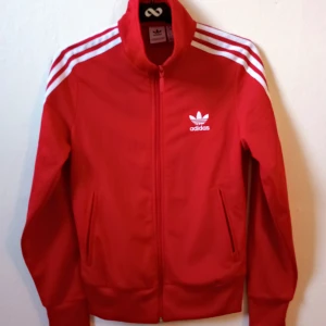 Röd träningsjacka från Adidas - Säljer en snygg röd träningsjacka från Adidas med de klassiska tre vita ränderna på ärmarna. Jackan har dragkedja och två fickor fram. Perfekt för träning eller som en sportig vardagslook. Den är i mycket bra skick och redo för nya äventyr! 🏃‍♀️