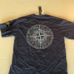 Mörkblå t-shirt från Stone Island - Säljer en snygg mörkblå t-shirt från Stone Island. Den har en stor logga på ryggen och en mindre logga på bröstet. Perfekt för en casual look eller att bära till vardags. T-shirten är i nytt skick och har korta ärmar.