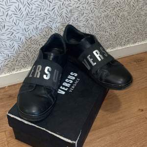 Snygga svarta sneakers från Versus Versace med breda remmar över foten där det står 'VERSUS'. Skorna är i skinn och har en stilren design som passar perfekt för både vardag och fest. De är i mycket bra skick och kommer med originalkartong.  Tveka inte på att fråga om ni har frågor pris kan diskuteras😊🙌🏽