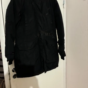 Svart parkajacka med pälsdetalj - Säljer en svart parkajacka med pälsdetalj på huvan. Jackan har långa ärmar och stora fickor framtill. Perfekt för kalla vinterdagar och höstpromenader. Den är i bra skick och håller dig varm och stilren.