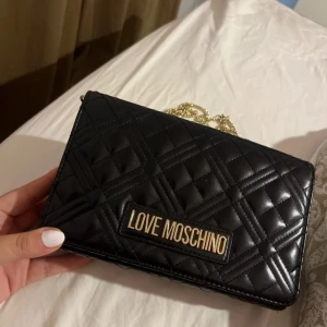 Svart quiltad axelväska från Love Moschino - Snygg svart axelväska från Love Moschino i quiltat mönster. Väskan har en elegant guldkedja som axelrem och en stilren logga framtill. Perfekt för både vardag och fest!