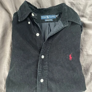 Svart manchester skjorta från Ralph Lauren - Säljer en snygg svart manchester skjorta från Ralph Lauren i storlek S. Den har en broderad röd logga på bröstet som ger en stilren touch. Mycket bra skick. Perfekt för både vardag och fest! Nypris 2k